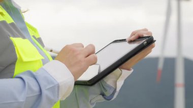 Yüksek görünürlüklü yelek giyen yetenekli bir profesyonel, yenilenebilir enerji alanındaki modern teknolojilerin rolünü vurgulayan bir rüzgar çiftliğinin ayrıntılı incelemesi sırasında tablet üzerindeki kritik verileri belgelemektedir.