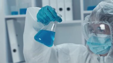 Bir laboratuvar ortamında, koruyucu ekipman içinde kendini adamış bir bilim adamı canlı mavi bir çözeltiye sahip cam şişeyi inceliyor, hassasiyeti ve güvenli deneyler için hayati önem taşıyor.