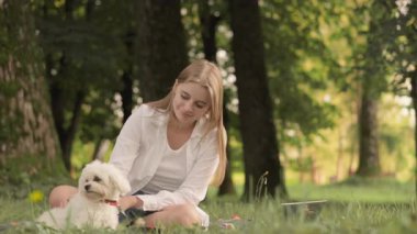 Huzurlu ve huzurlu bir park ortamında, neşeli genç bir kadın yemyeşil çimenlerin üzerinde oturuyor, neşeli köpeğini neşeyle okşuyor, güzel bir arkadaşlık anının ve doğayla olan bağının tadını çıkarıyor.