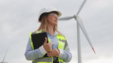 Parlak yansıtıcı giysiler giymiş bir kadın mühendis, yenilenebilir enerji alanındaki yenilik ve adanmışlığın özünü temsil eden güçlü bir rüzgar türbininin yanında güvenle duruyor.