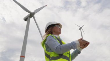 Güvenlik teçhizatında kararlı bir kadın mühendis arka planda türbin alanı olan bir rüzgar türbini modelini inceleyerek yenilenebilir enerji ve sürdürülebilirliğe olan bağlılığı sembolize ediyor.