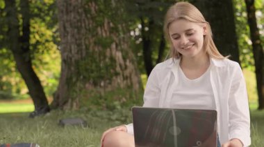 Açık sarı saçlı genç bir kadın laptopunu açık havada, doğa ve uzun ağaçlarla çevrili huzurlu bir parkta kullanırken gülümsüyor, rahatlama ve üretkenliğin özünü somutlaştırıyor..