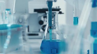 Mavi eldivenli bir bilim adamının deney tüpleri ve mataralara pipet kullanarak mavi bir sıvıyı ustalıkla aktardığını gösteren bir dizi laboratuvar görüntüsü bilimsel deneylerdeki hassasiyeti vurguluyor..