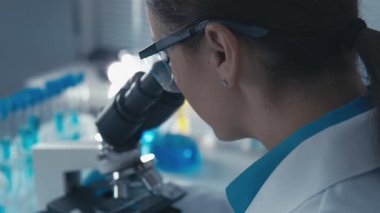 Tutkulu bir kadın bilim adamı, mavi cam ve laboratuvar ekipmanları ile çevrili mikroskopla örnekleri inceleyerek çığır açan araştırmasına ve biyoteknoloji yeniliklerine odaklanıyor.