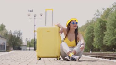 Şık yaz kıyafetleri içinde neşeli genç bir kadın yoğun bir tren istasyonunda parlak sarı bavuluyla oturmuş macera ve mutluluk dolu heyecanlı bir tatil için sabırsızlıkla bekliyor.