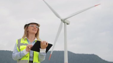 Kasklı ve yansıtıcı yelek giyen profesyonel bir kadın mühendis, rüzgâr türbininin önünde dikilirken bir tablet üzerindeki verileri gözden geçirir. Sürdürülebilirlik teknolojisini iş başında gösterir..