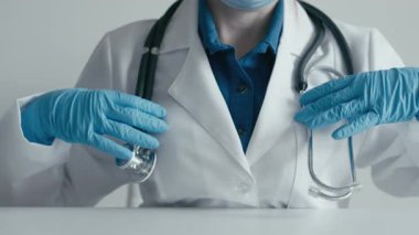 Beyaz önlüklü ve mavi eldivenli kendini adamış bir tıp uzmanı steteskop kullanarak hastalarla yapılan tipik sağlık muayenelerinin tipik klinik atmosferini vurguluyor.