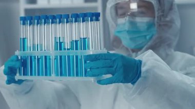 Koruyucu giysi giyen bir bilim adamı steril bir laboratuvar ortamında canlı mavi çözümlerle dolu bir test tüpü rafını inceliyor..