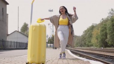 Renkli yaz kıyafetleri içinde hayat dolu genç bir kadın hareketli bir tren istasyonunda bir bavulla neşeli bir şekilde geziniyor, heyecanlı ve manzaralı bir yolculuk için hazır hissediyor.