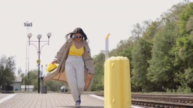 Parlak yaz kıyafetleri içinde genç bir kadın, yoğun bir tren istasyonunda, maceracı ruhunu ve seyahat heyecanını simgeleyen büyük bir bavulla güvenle yürüyor.