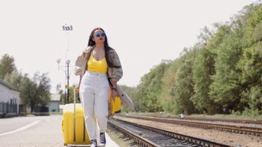 Parlak yaz kıyafetleri içinde şık bir kadın, seyahat ve macera ruhunu sergileyen neşeli sarı bir bavulla dolu, yoğun bir tren istasyonunda güvenle yürüyor.