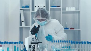Steril bir laboratuvarda, koruyucu ekipman içinde kendini adamış bir bilim adamı test tüplerini inceler ve mikroskop kullanır. Gelişmiş araştırma tekniklerini göstererek bilimsel keşifleri engeller.