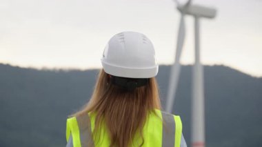 Sert şapka ve yansıtıcı yelek giymiş bir kadın mühendis, yenilenebilir enerjiye ve sürdürülebilir gelişime olan bağlılığını güzel bir doğal ortamda sergileyerek büyük bir rüzgar türbini inceliyor.