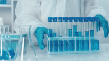 Koruyucu giysi alanında çalışan kendini adamış bir bilim adamı test tüplerindeki canlı mavi çözümleri analiz ediyor. Laboratuvar araştırmalarının özünü ve günümüz bilimsel ortamındaki sıkı güvenlik protokollerini somutlaştırıyor.