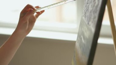 Bir el, tuval üzerinde çalışırken boya fırçasını nazikçe tutarken çekilmiş yakın plan bir fotoğraf. İçerideki ortam, doğal ışığın sanatsal süreci hafifçe aydınlattığı gibi.. 