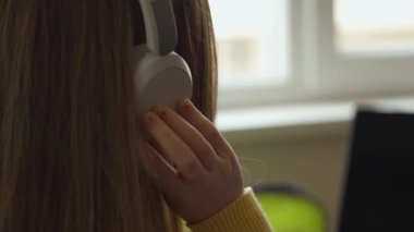 Kulaklıkta odak noktası olan, kulaklık takan ve el kulaklığın üzerinde dinlenen birinin yakın çekimi. Arka plan parlak bir iç mekan olduğunu gösteriyor, muhtemelen bir ofis..