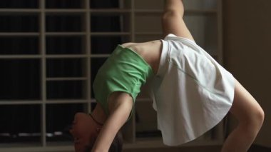 Bir kadın içeride gücünü ve esnekliğini sergileyerek ters köşe yapıyor. O, yeşil bir üst ve beyaz bir etek giymektedir. Arkaplanı koyu tonlar doldurur.