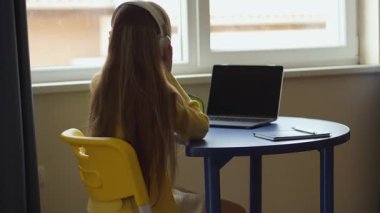 Sarı kazaklı ve kulaklıklı birinin mavi bir masada, pencerenin yanında dizüstü bilgisayar kullanırken görüntüsü. Ev ortamında dizüstü bilgisayarın yanında bir not defteri ve kalem var..