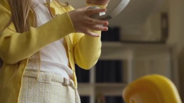 Yakın plan çekimde, üzerinde güneşli sarı bir hırka, beyaz bir bluz ve desenli bir etek olan, fark edilemez bir nesne tutan bir kişi görülüyor. Kapalı alan ayarları rahat, arkaplanda bir kitaplık var..