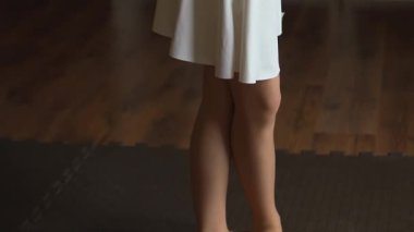 Dansçıların bacakları, beyaz bir elbise giyen yakın çekim görüntüsüyle tasvir edilir. Dansçı ahşap ve siyah bir zeminde duruyor. Işıklandırma minimalist ama zarif bir atmosfer yaratıyor.