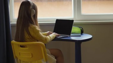 Genç bir kız masada oturuyor, dizüstü bilgisayar kullanıyor ve kulaklık takıyor. Pencerenin yanındaki kapalı bir mekanda, bilgisayarında öğrenme ya da eğlence ile meşgul. Sahne parlak ve odaklı.