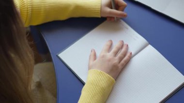 Bir kişi çizelge kağıdıyla deftere yazı yazıyor. Sarı bir kazak giyiyorlar ve mavi bir masaya yazı yazıyorlar. Kalemi tutan ele ve açık olan deftere odaklanıyoruz..