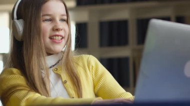 Kulaklıklı genç bir kız bilgisayarıyla mutlu bir şekilde meşgul oluyor, muhtemelen online bir derse katılıyor ya da bir proje üzerinde çalışıyor. Arka plan ev içi çalışma ortamının rahat olduğunu gösteriyor..