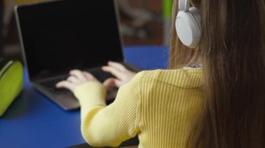 Kulaklıklı bir insan dizüstü bilgisayarda çalışıyor. Mavi bir masada solda bir nesneyle oturuyorlar. Kişi sarı bluzlu ve laptop ekranı boş..