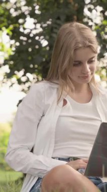 Genç bir kadın, çalışmalarını bir dizüstü bilgisayara derin bir şekilde daldırmış, rahat bir şekilde yeşil çimlerin üzerinde, canlı yeşilliklerle çevrili ve ılık, yumuşak güneş ışığıyla güneşleniyor.