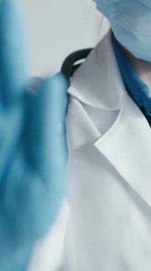 Beyaz önlüklü ve eldivenli bir sağlık çalışanı, cerrahi maske takarken dikkat göstererek, sağlık ortamlarındaki herkesin sağlığını korumak için güvenlik protokollerinin önemini vurgular.