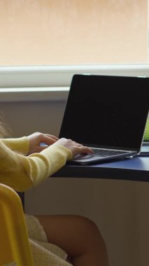 Genç bir kız masada oturuyor, dizüstü bilgisayar kullanıyor ve kulaklık takıyor. Pencerenin yanındaki kapalı bir mekanda, bilgisayarında öğrenme ya da eğlence ile meşgul. Sahne parlak ve odaklı.