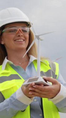 Güvenlik donanımlı kendinden emin bir kadın mühendis, yenilik ve sürdürülebilirliği temsil eden bulutlu gökyüzünün altında yenilenebilir enerji alanında detaylı bir rüzgar türbini modeli sergiliyor.