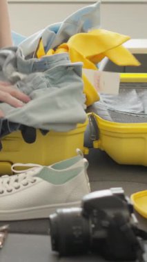 Kişi titizlikle parlak sarı bir bavulu giysi ve seyahat eşyalarıyla dolduruyor. Etrafı bitkilerle çevrili ve tatil hazırlığı için ideal bir ortam.