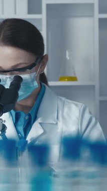 Kendini adamış bir kadın bilim adamı, renkli test tüpleri ve temel laboratuvar ekipmanlarıyla çevrili mikroskopla örnekleri analiz ederek, alanındaki araştırma ve yeniliklerin önemini vurguluyor.