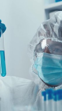 Koruyucu giysi giyen bir bilim adamı modern bir laboratuvarda parlak mavi sıvıyla bir test tüpünü inceleyerek araştırma, keşif ve teknolojide ilerlemenin hayati önemini vurguluyor.