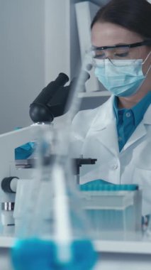 Laboratuvar önlüğü giymiş odaklanmış bir bilim adamı mikroskop kullanarak renkli test tüpleriyle çevrelenmiş örnekleri inceliyor, çağdaş bilimsel araştırmalarda hassasiyet ve yenilik sergiliyor..