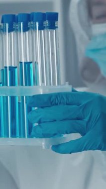 Koruyucu giysi giyen bir bilim adamı steril bir laboratuvar ortamında canlı mavi çözümlerle dolu bir test tüpü rafını inceliyor..