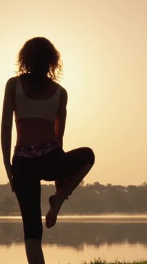 Sabah ışığında, bir kadın huzur ve uyumu somutlaştıran sakin bir gölün yanında yoga yapıyor, doğayla derin bir bağ kuruyor, seyircilere hareketleriyle ve sakin tavırlarıyla ilham veriyor.