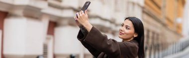 happy brunette woman taking selfie on mobile phone on blurred street in prague, banner clipart