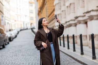 Çapraz vücutlu şık esmer kadın Prag 'da cep telefonundan selfie çekiyor.