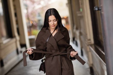 young brunette woman tying coat belt on urban street in prague clipart