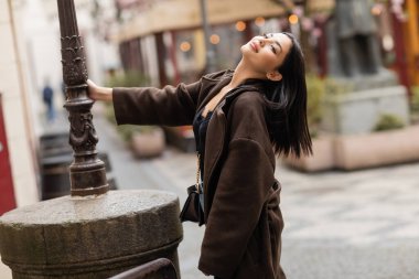 sensual brunette woman in brown autumn coat posing near lamppost in prague clipart