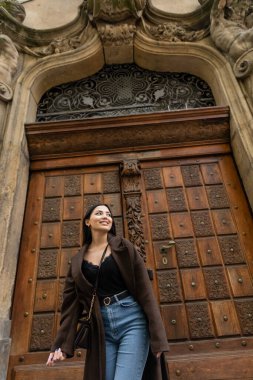 Prag 'da oyulmuş ahşap kapının yanında gülümseyen kot pantolonlu esmer kadının düşük açılı görüntüsü. 