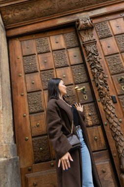 young brunette woman in brown coat opening wooden carved door in prague clipart