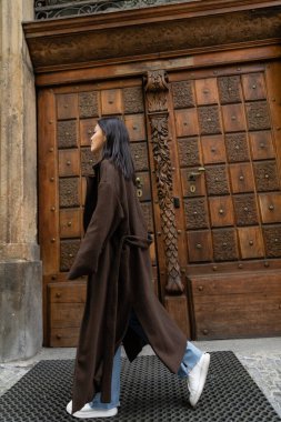 side view of brunette woman in stylish coat walking near carved wooden door on street in prague clipart