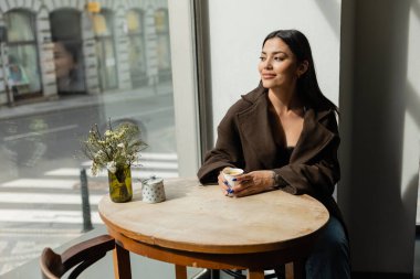 happy woman looking at urban street while sitting near window in cafe in prague clipart