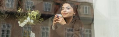 smiling woman with disposable cup looking through window in prague cafe, banner clipart