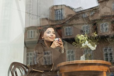 young brunette woman in coat drinking coffee from paper cup near window in prague cafe clipart