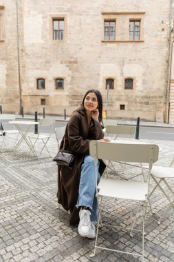 Trendy young woman in coat looking away near paper cup with coffee in outdoor cafe in Prague  clipart