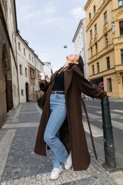 Side view of joyful woman in coat holding paper cup on street in Prague  clipart